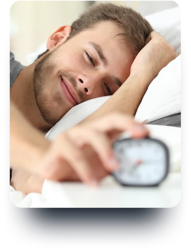 A young man, looking relaxed and lying on the bed, about to stop an alarm clock.